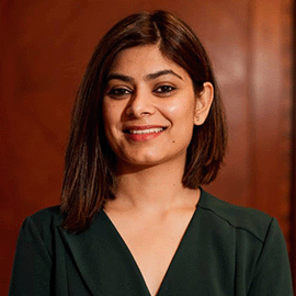 A smiling woman standing in front of a wooden background looks into the camera.